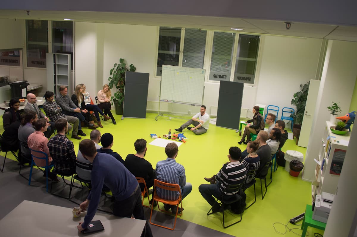 A group of people sitting in a half circle on chairs while a workshop host is sitting on a floor in the middle with random sortment of items next to them