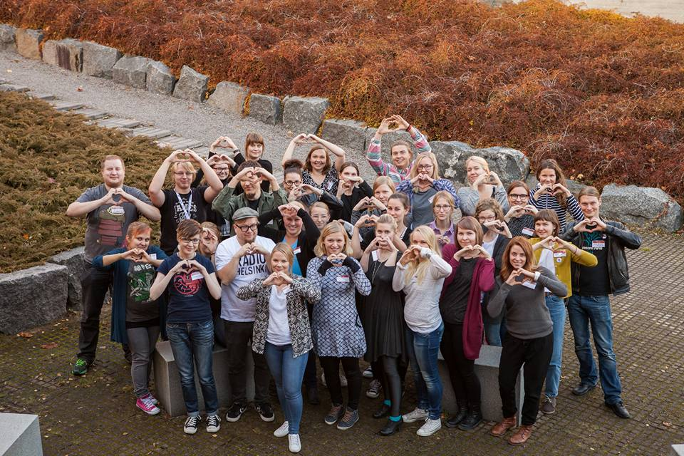 A group photo taken outside on a concrete yard. A group of people standing close to each other, all showing a heart sign with their hands.