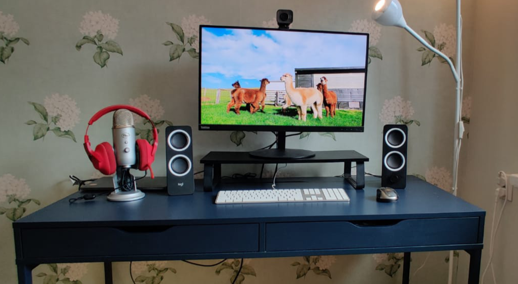 A desk with closed macbook connected to a display, white keyboard and black/grey mouse, small black standing speakers and a Yeti microphone with red headphones
