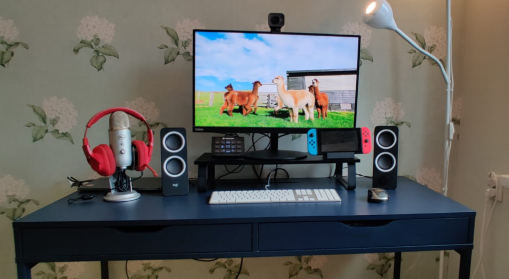 A desk with closed macbook connected to a display, white keyboard and black/grey mouse, small black standing speakers and a Yeti microphone with red headphones. A Nintendo Switch on one side of the display and Stream Deck on another