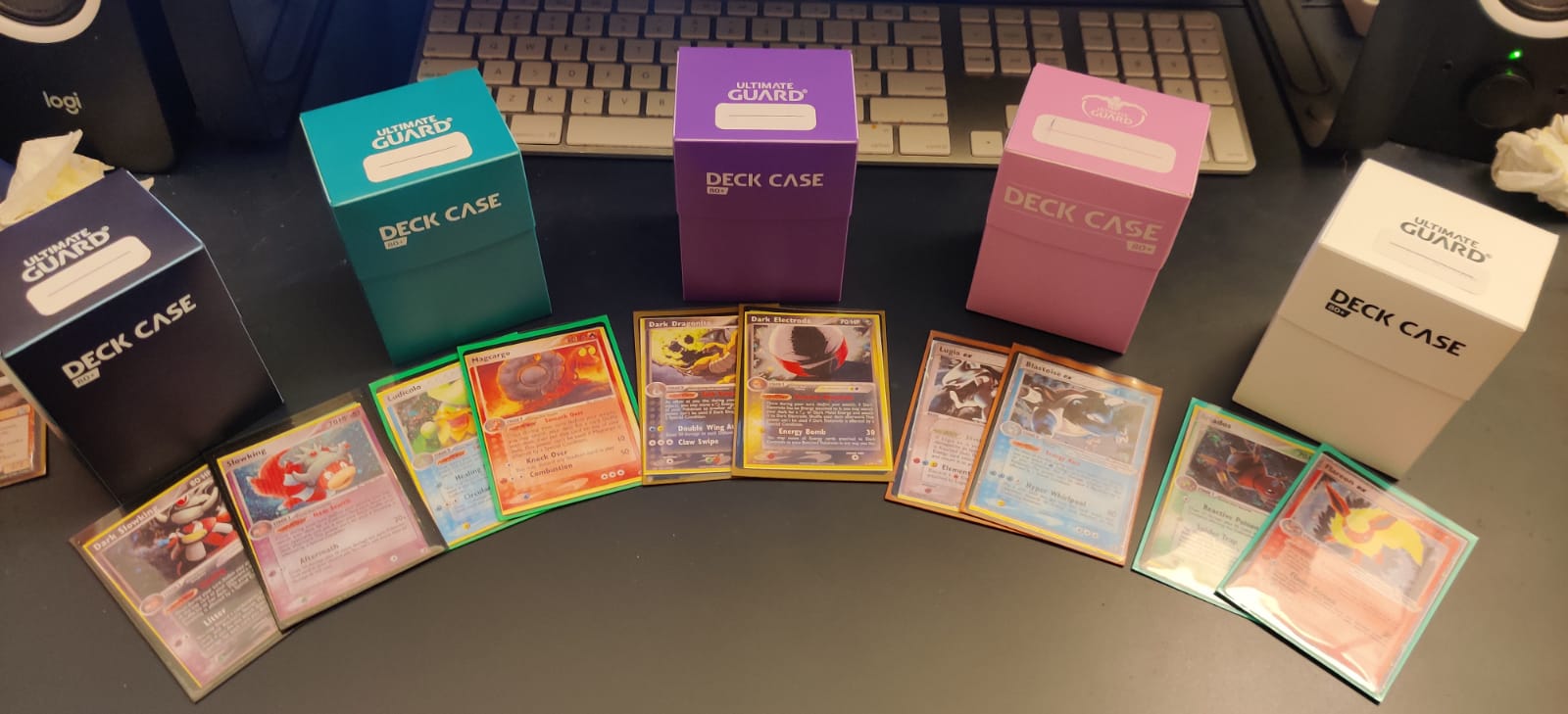 Five different color deck boxes in a fan formation and in front of each there are two Pokemon cards unique to decks that live in those boxes
