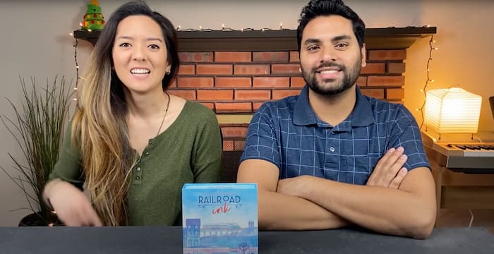 A woman and a man sitting at a table, looking into the camera with Railroad Ink board game box in front of them