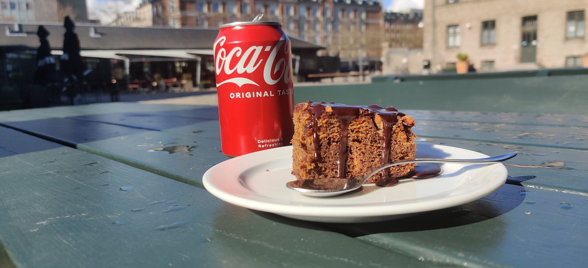 A can of coke and a piece of chocolate cake, taken outside on a sunny afternoon day