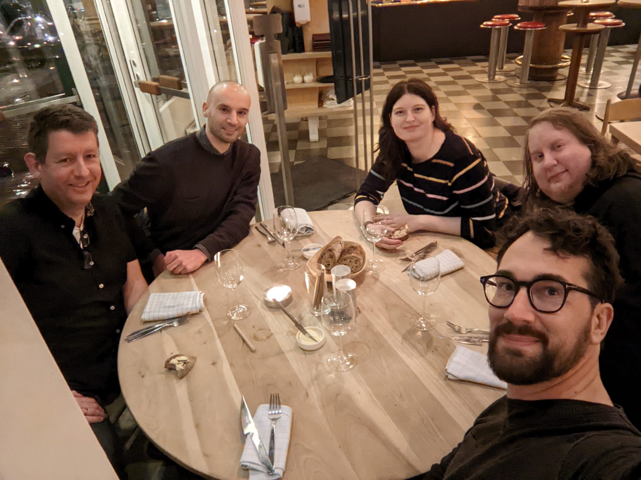 Five people sitting around a round table in a restaurant, waiting for food to arrive, all smiling and looking into the camera.