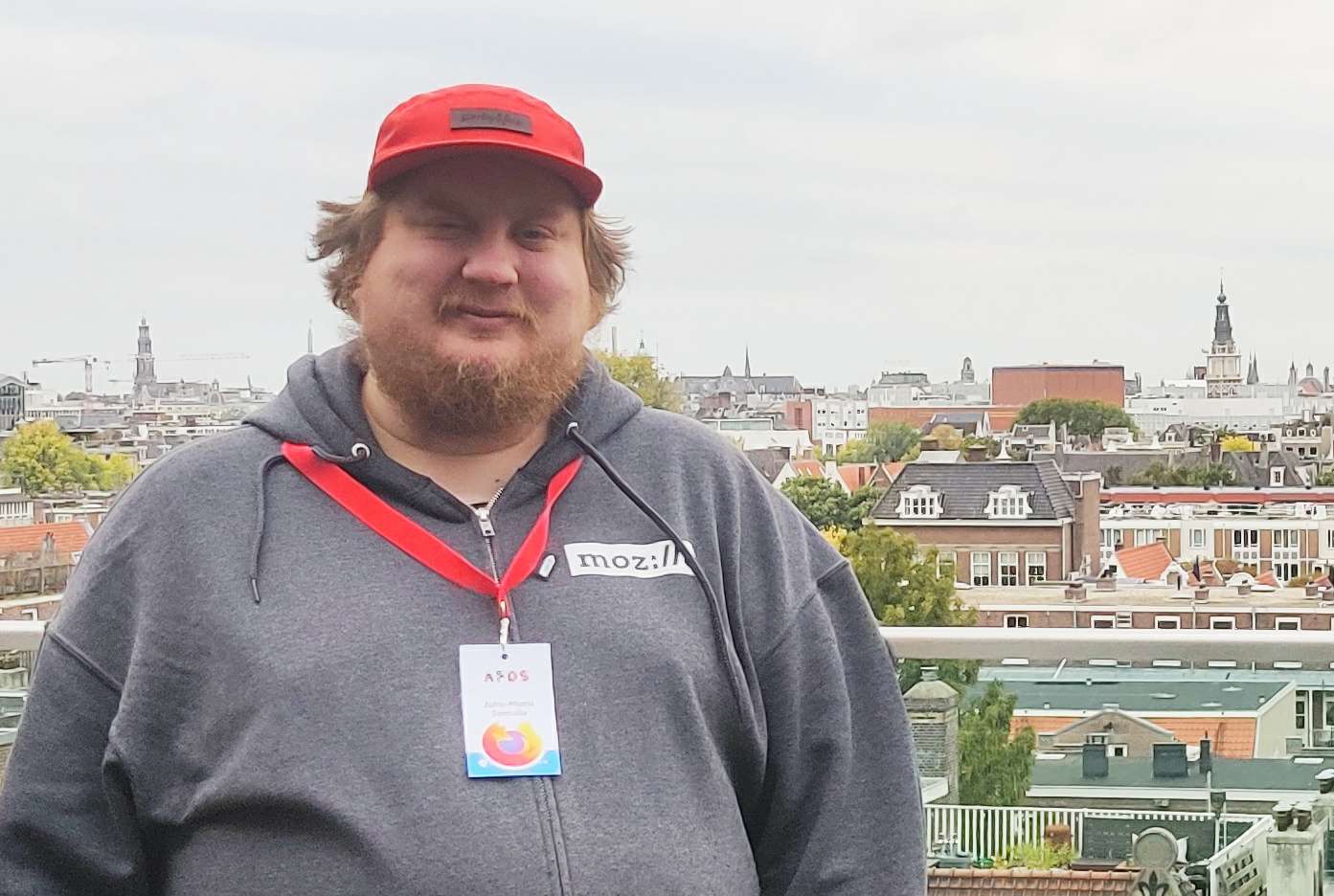  Me standing outside, wearing a red cap and grey hoodie with Mozilla logo. I have  conference badge with a Firefox sticker hanging from my neck. Behind me, there are the rooftops of Amsterdam.