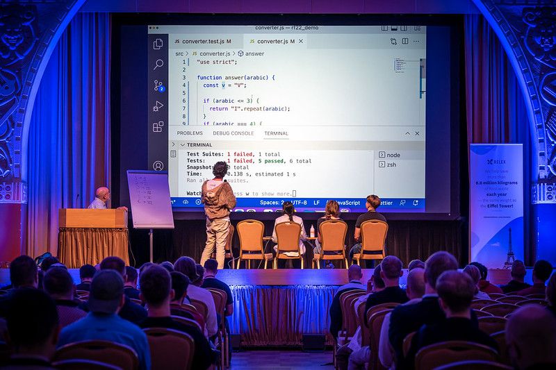 Three people sitting on chairs in the stage, their backs towards the audience. Another person stands next to them, back to audience. All of them are looking into the screen at the end of the hall. There's Python code on the screeen.