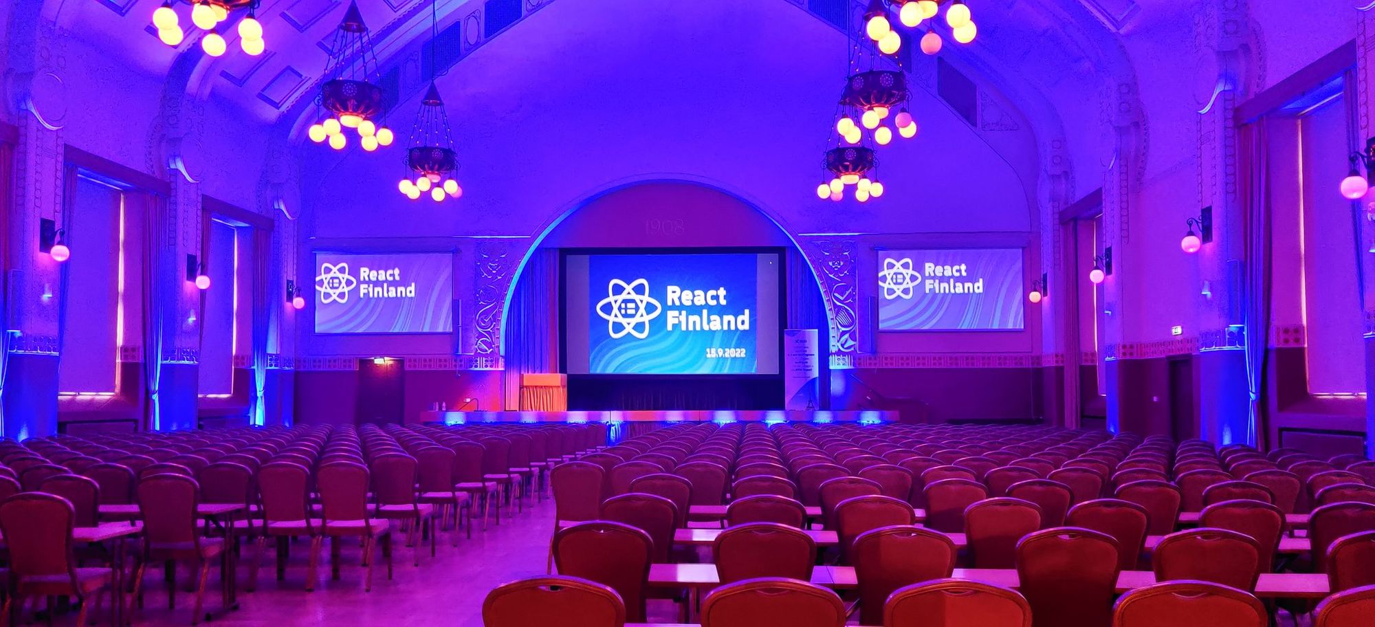 An empty conference hall with purple ambience coloring, hundreds of chairs and a React Finland logo on three big screens at the end of the hall