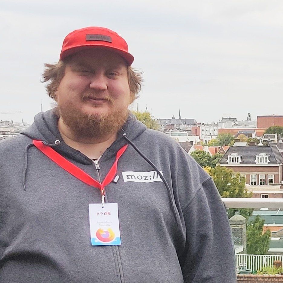 Juhis wearing a red cap, grey hoodie with Mozilla hoodie and a conference bad, standing outside with rooftops of Amsterdam in the background