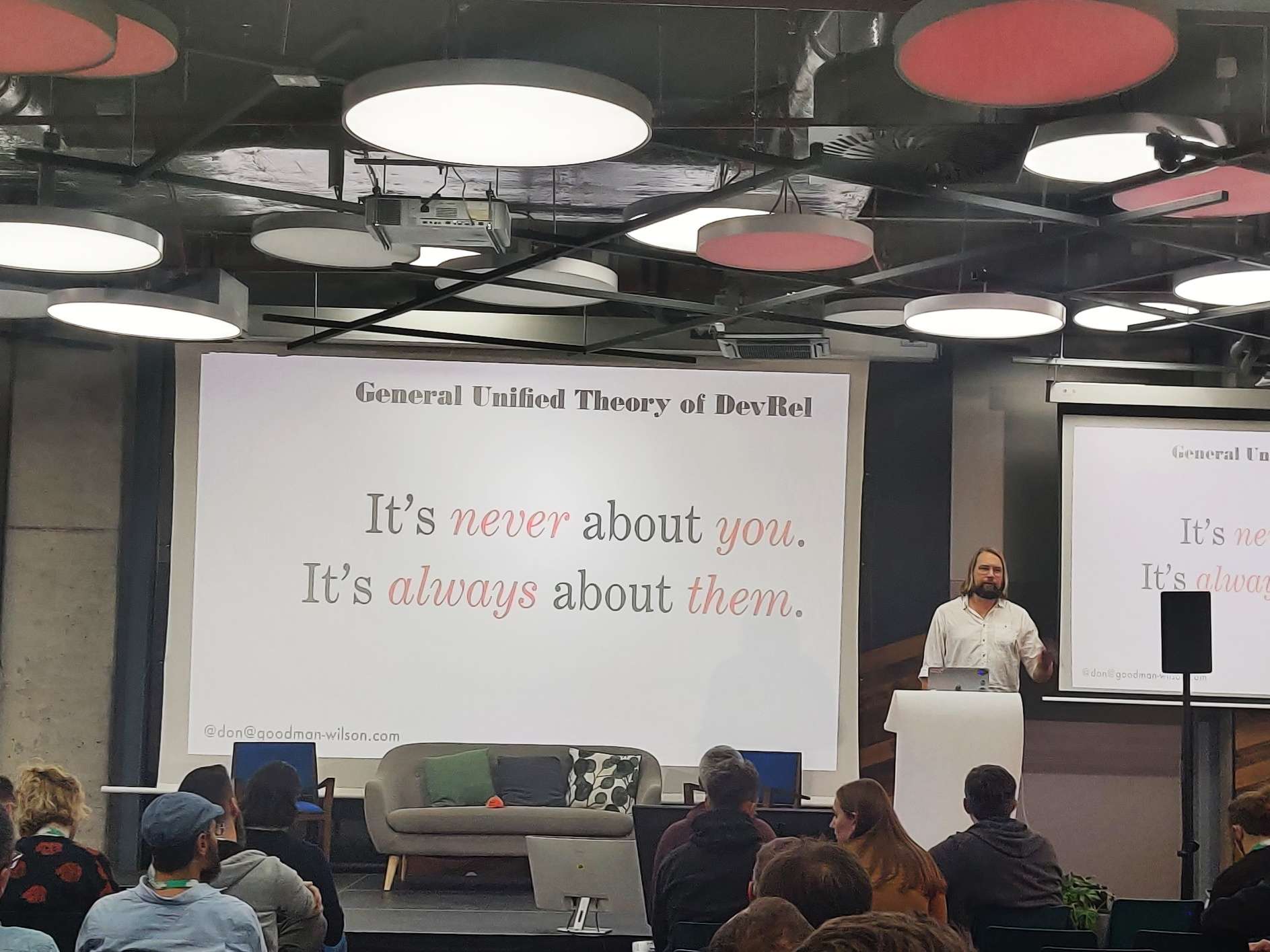 Don standing on stage wearing a white shirt. A slide project next to him says "It's never about you. It's always about them."