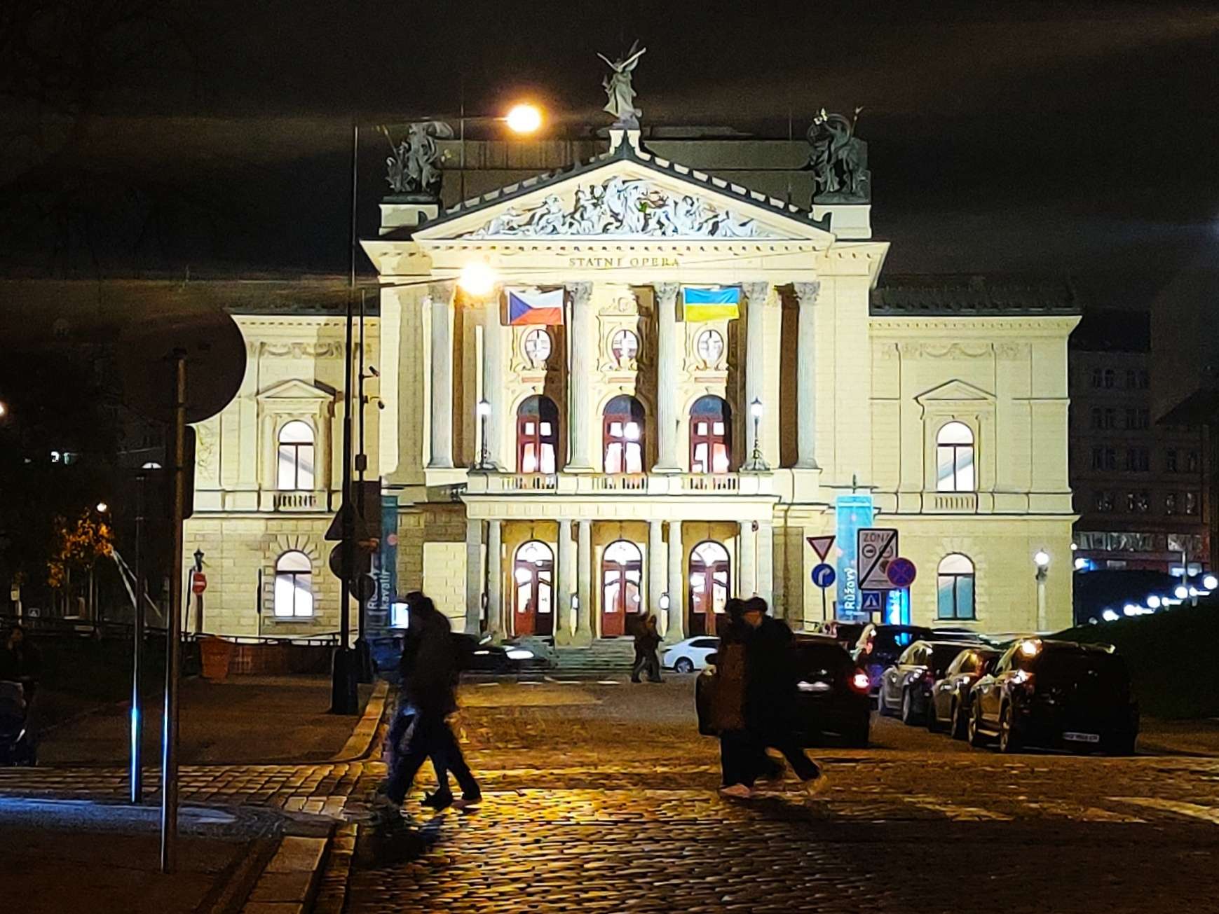 Prague's state opera house in the evening, lit up with lights. Czech and Ukrainian flags hanging on the outside.