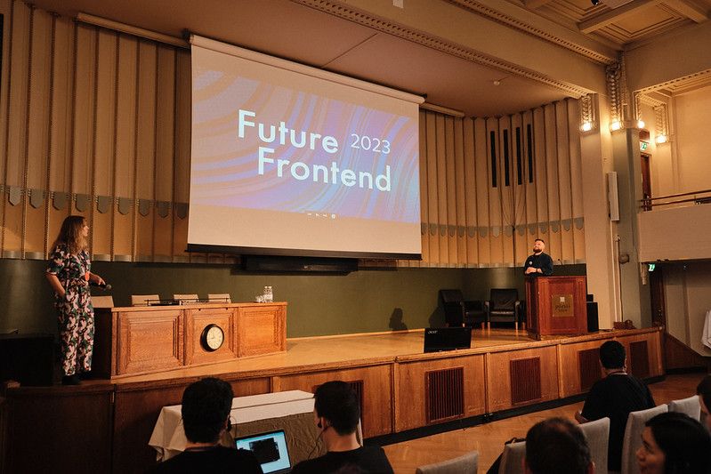 Future Frontend logo on a large screen at a stage with a woman and man standing on opposite sides of the stage, speaking to the audience.