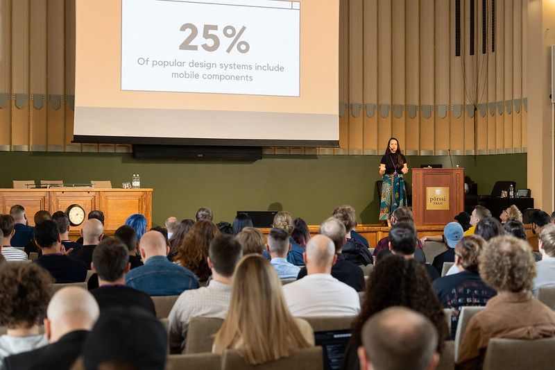 Thaís standing on a stage with a slide on the background that reads '25% of popular design systems include mobile components'