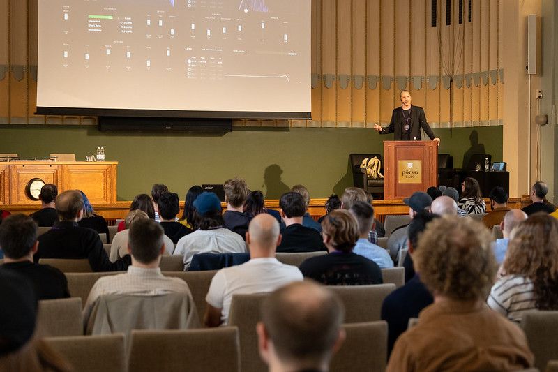 Tero standing behind a speaker booth on the stage with a slide barely visible on the background showing lots of slider toggles. On the foreground, there's a bunch of people sitting in the conference hall, listening to Tero speak.