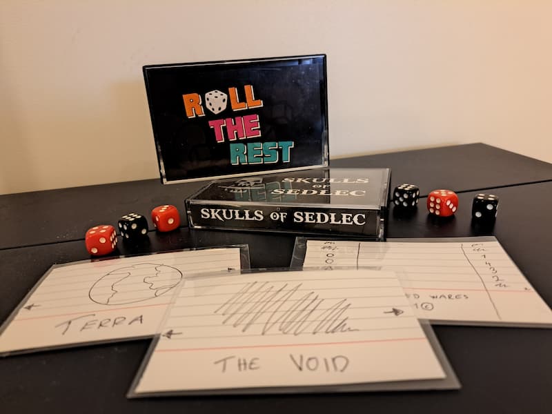 Couple of hand-drawn playing cards on index papers inside transparent sleeves, a few dice scattered around and two C cassette boxes, one with Roll the Rest text logo in front and another one with Skulls of Sedlec text logo in the spine. 
