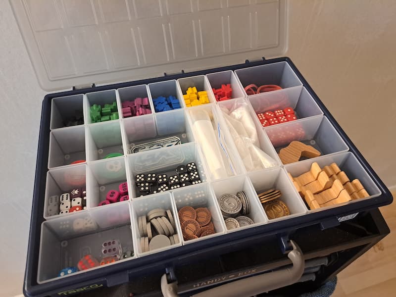 A box with a lot of small compartments filled with variety of colorful board game tokens, meeples and dice. 