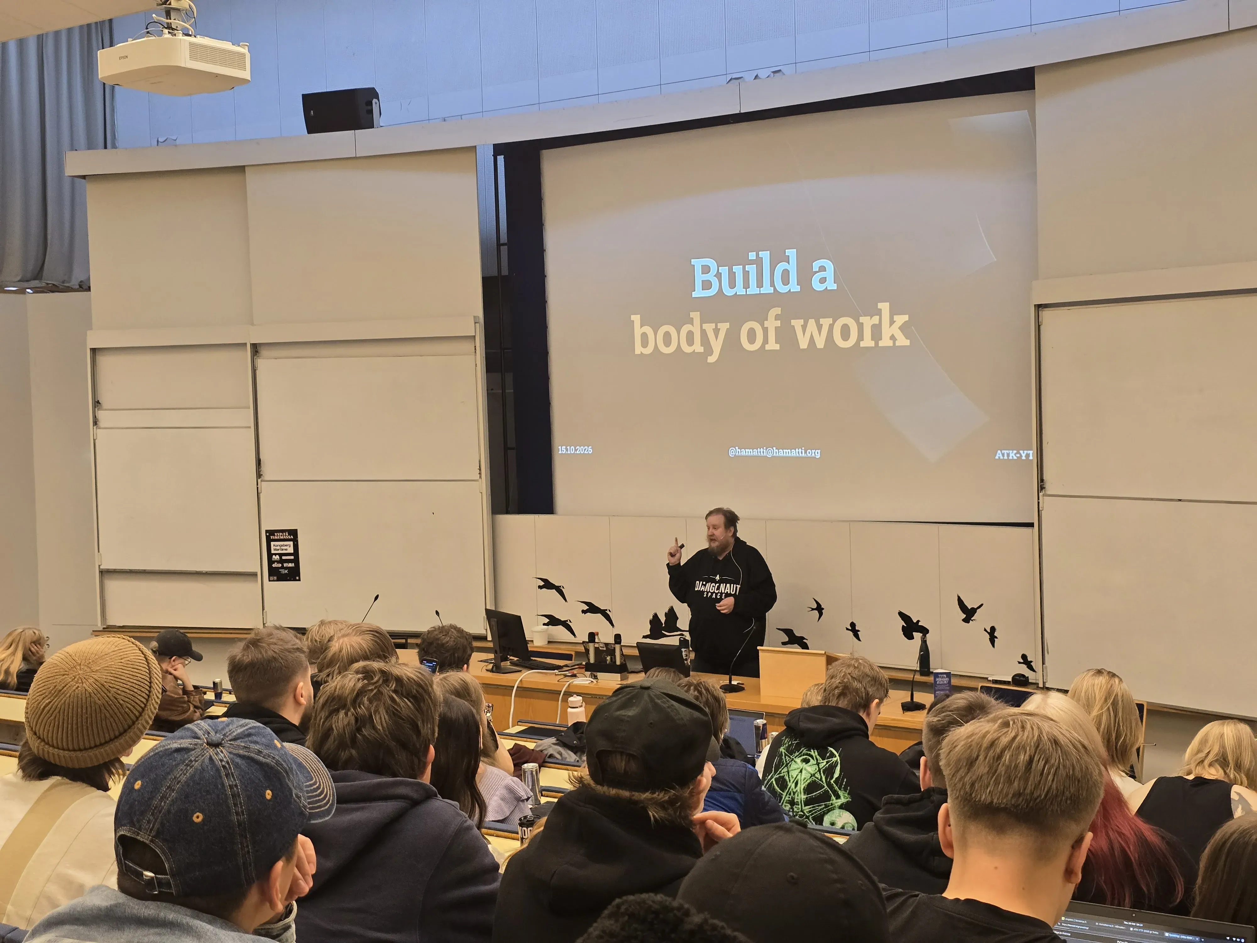 Juhis speaking in a lecture hall to a room full of students. On the screen, there’s a slide with text: Build a body
    of work. 