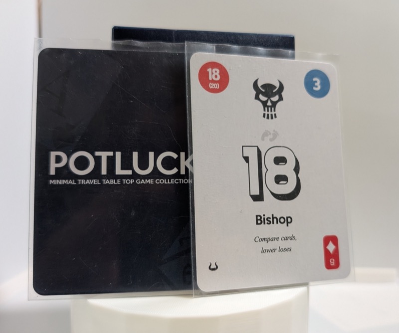 Two sleeved playing cards. On the left, the card is showing its dark blue black with text Potluck: Minimal Travel Table Top Game Collection. On the right, the card has large number 18 in the middle, a diamond 5 traditional card suit, numbers 18 and 3 in top corners, a single bull horn in the bottom left corner and a skull center top. Center bottom, a text Bishop: Compare cards, lower loses. 