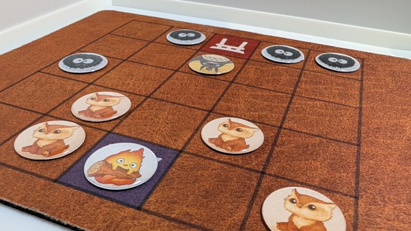 A mousepad with a brown background and a printed 5 by 5 grid on top. Circular tokens with art of Calcifer fire demon, cute owlbears, Totoro and dust bunnies are scattered around the grid. In the center position on two opposite ends of the grid are tori gates in blue and red blackgrounds. 