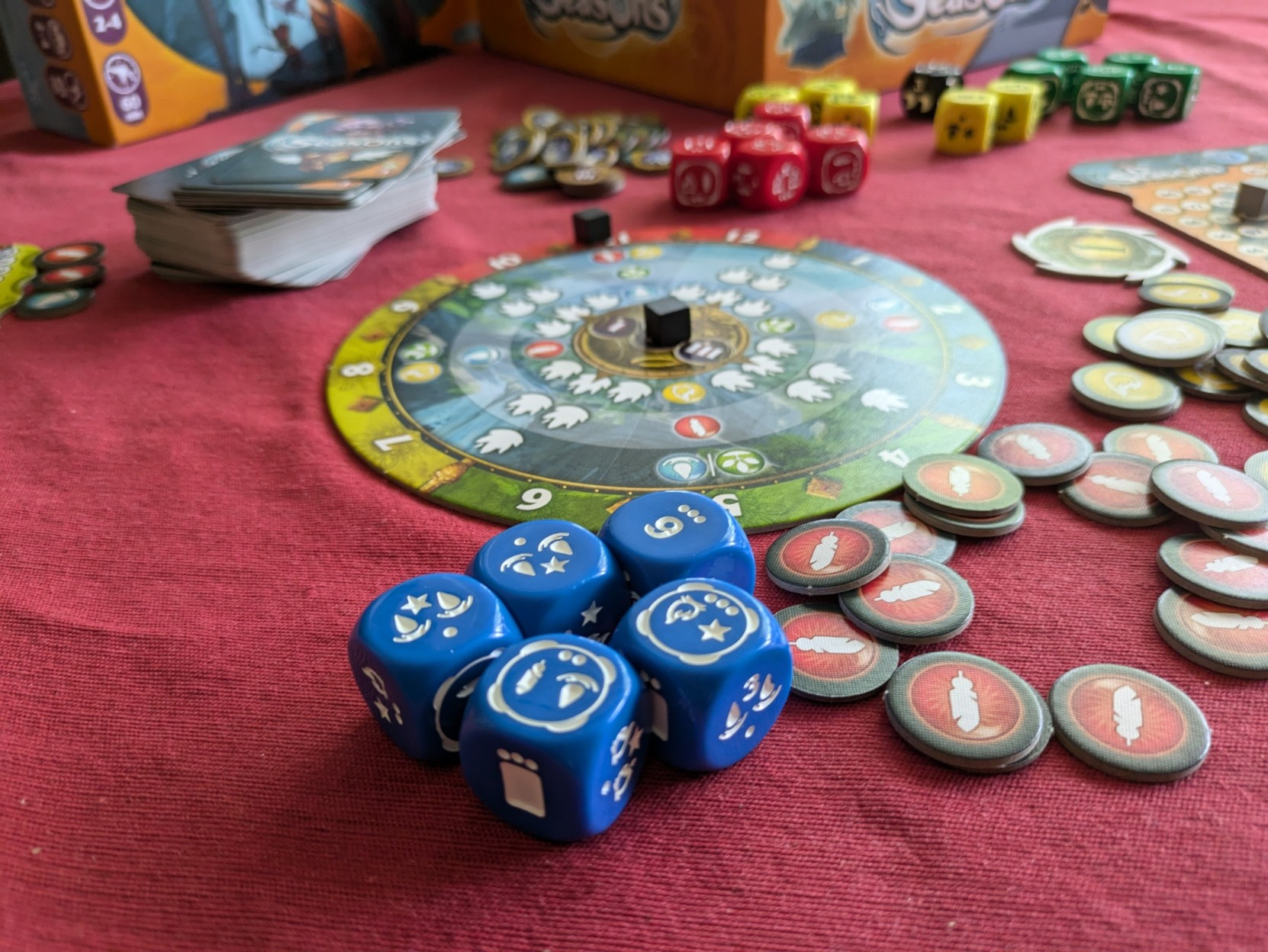 Bunch of board game components from Seasons spread on the table. In front, five blue dice with different markings, behind them a circle game board and scattered around some round energy tokens. 