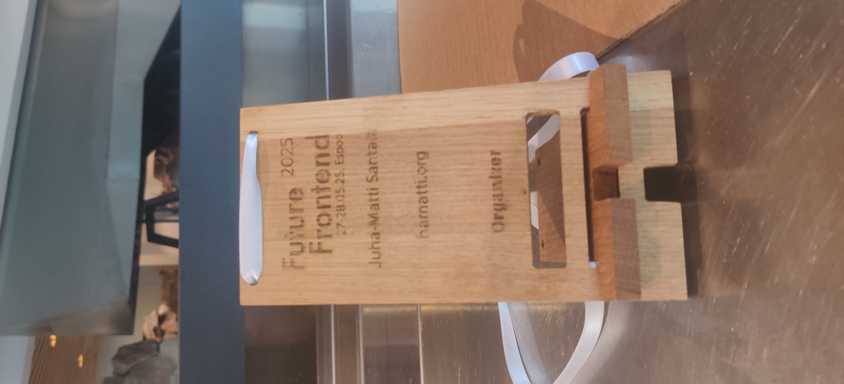 A wooden phone stand with engraving of Future Frontend 2025 and Juha-Matti Santala / organizer. A white lanyard goes through two holes at the top, making it function as a conference badge as well.