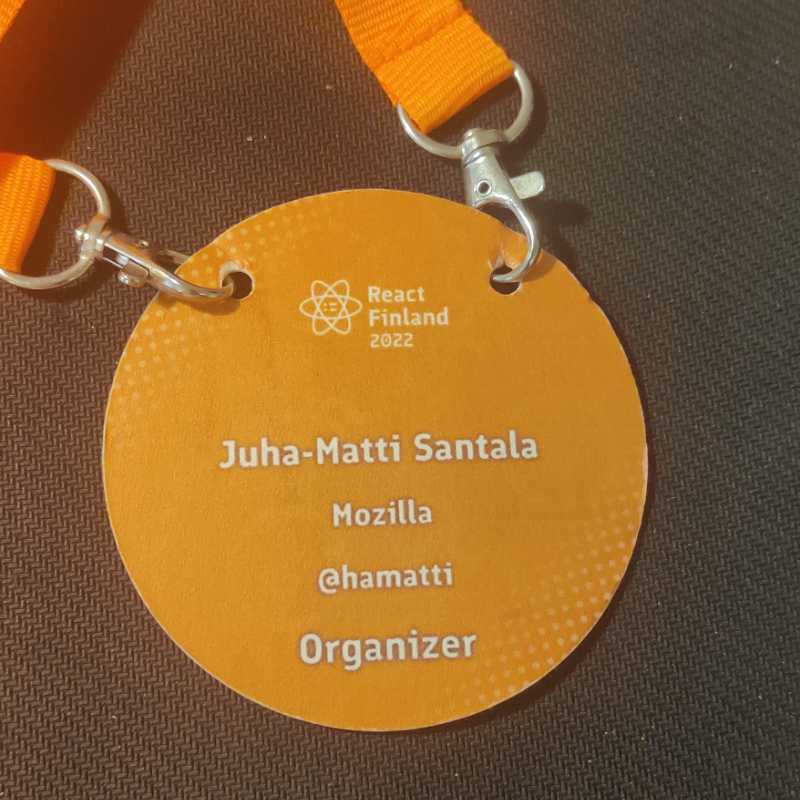A round, bright orange conference badge for React Finland 2022 with the event logo, my name, company name, Twitter handle and role “Organizer”. 