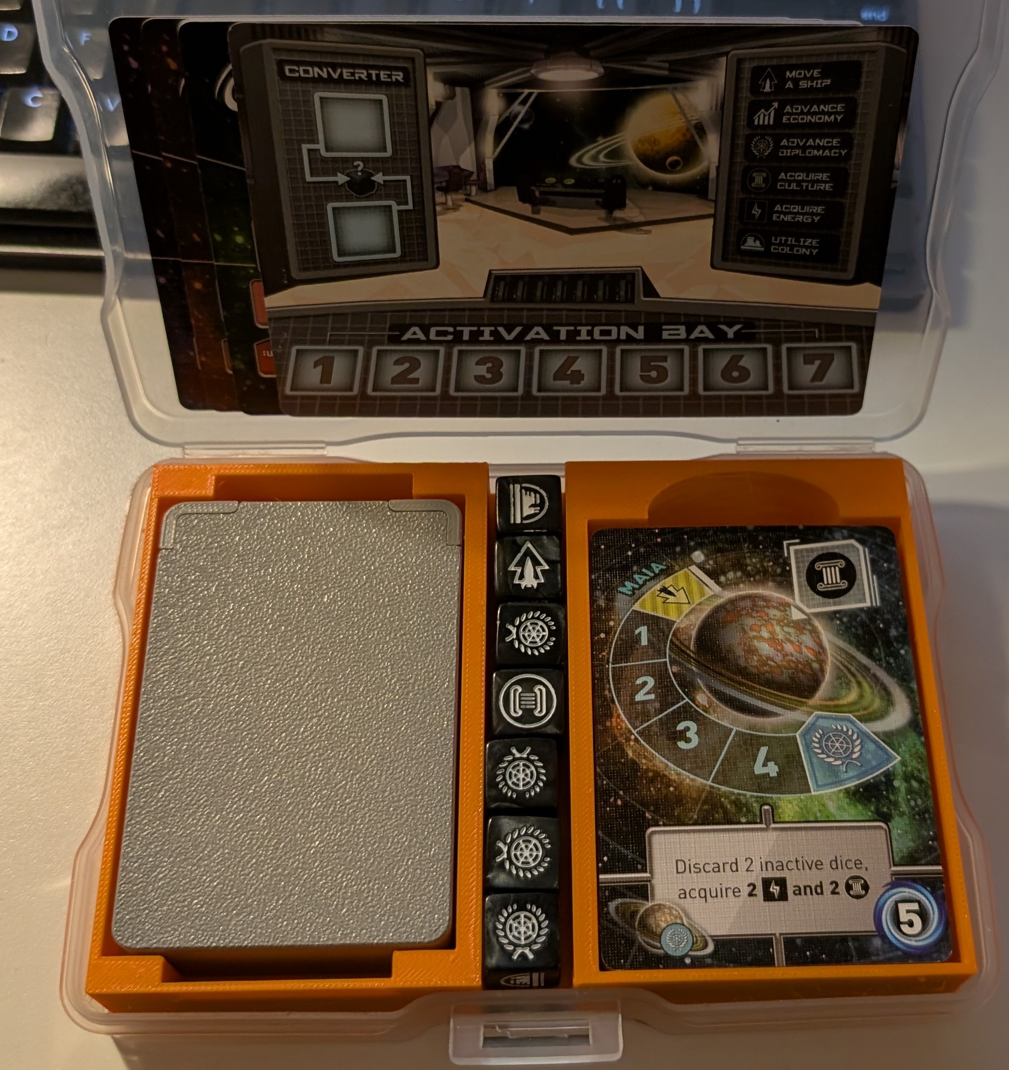 An orange 3D printed insert for Tiny Epic Galaxies inside a transparent plastic photo caddy box. On the left side area of the insert, there’s a smaller, grey box. 