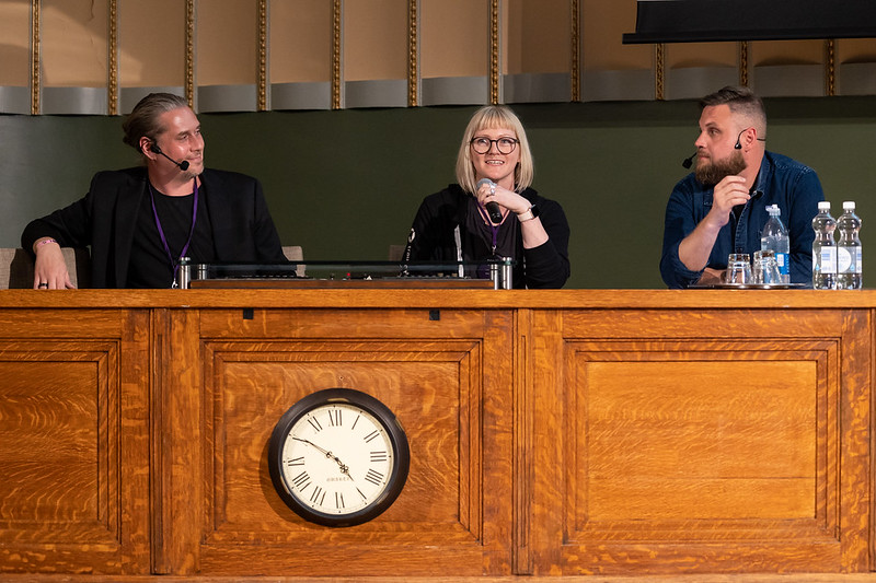 Tero, Stephanie and Jani sitting behind a wooden desk that has a clock in the front face. Stephanie is holding a microphone while Tero and Jani are wearing headset mics. 