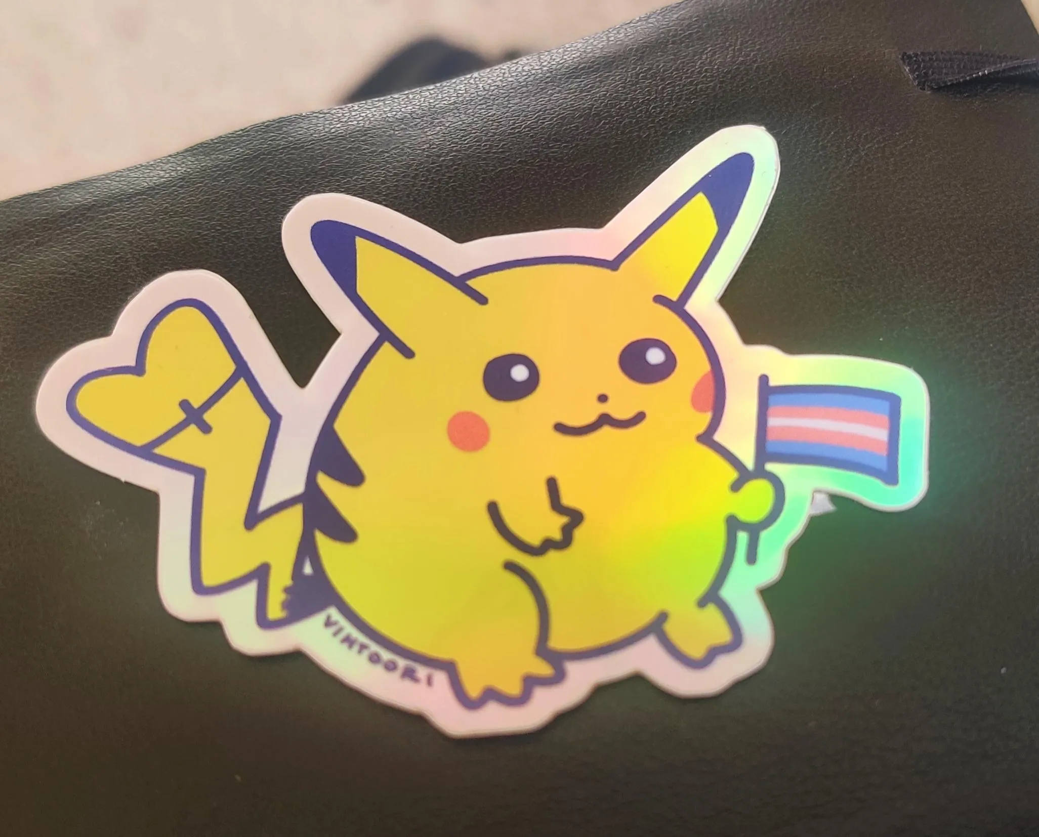 A sticker of Pikachu holding a trans flag and a scarred/stitched end of tail with a heart shape. 