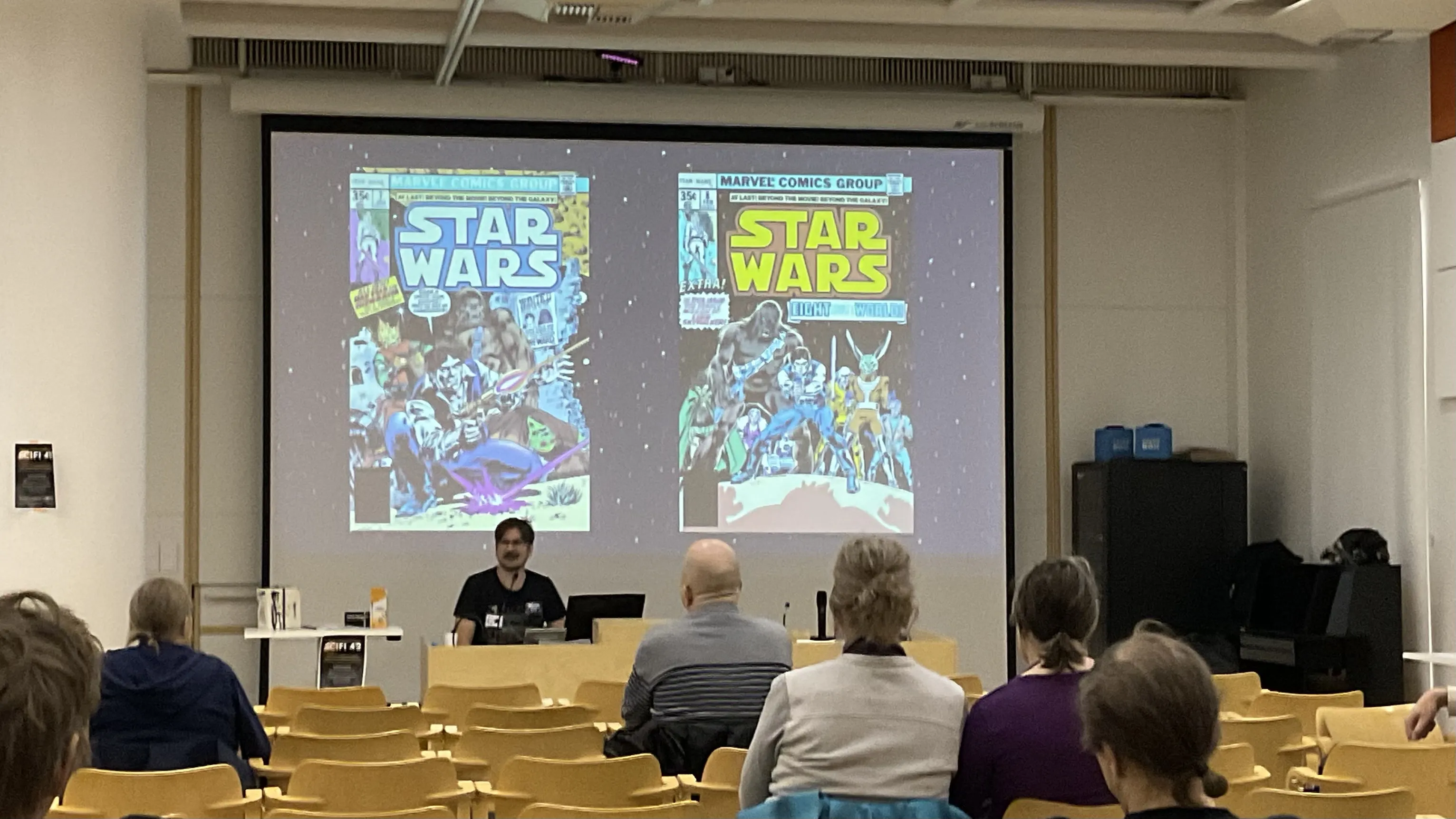 Juha sitting in front of a room, presenting with two Star Wars comics covers on a screen behind him. There are a handful of people visible in the audience.
