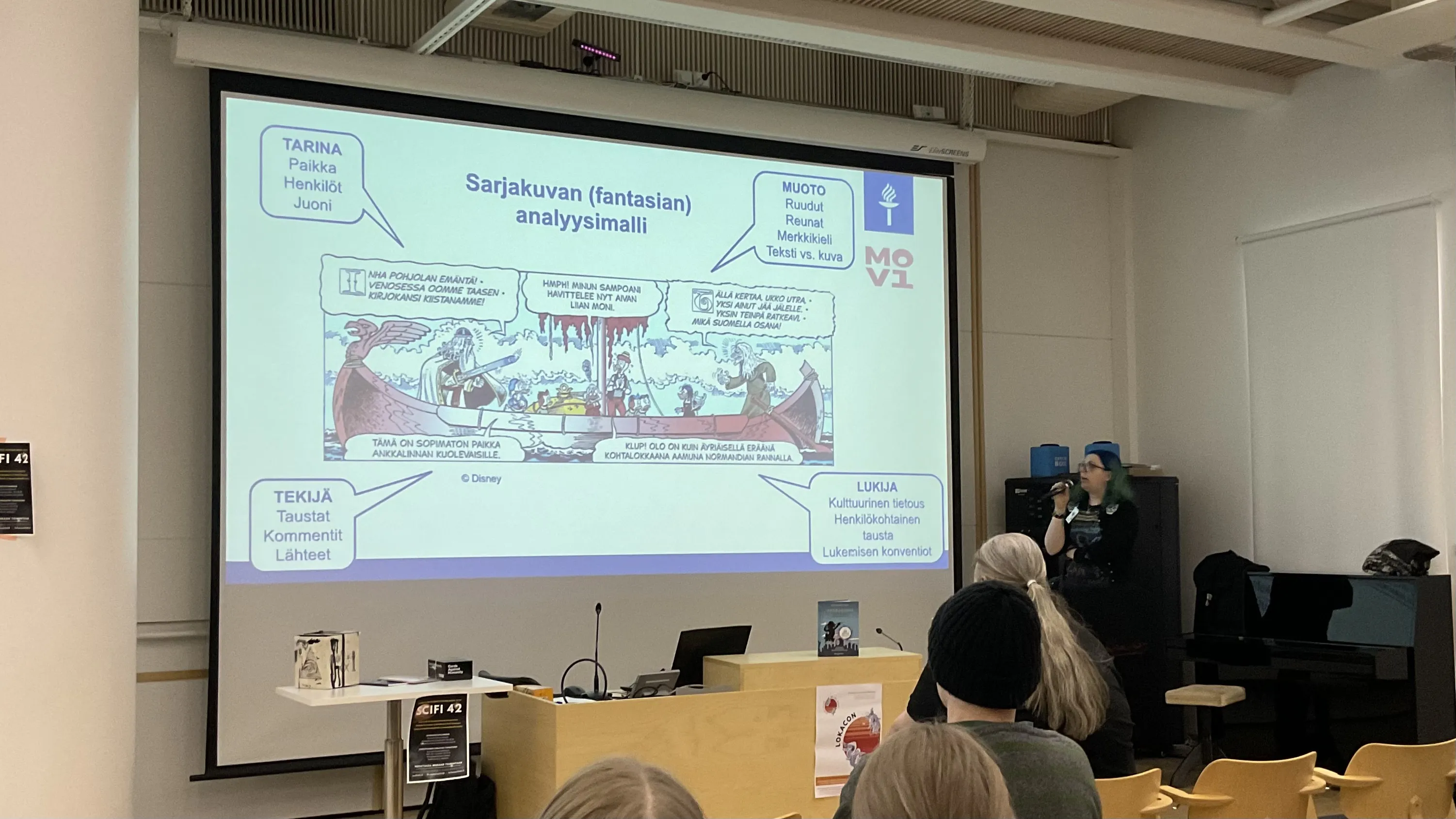 Katja speaking with a Don Rosa Donald Duck comic on the screen and text in Finnish around it explaining how to analyse fantasy comics.