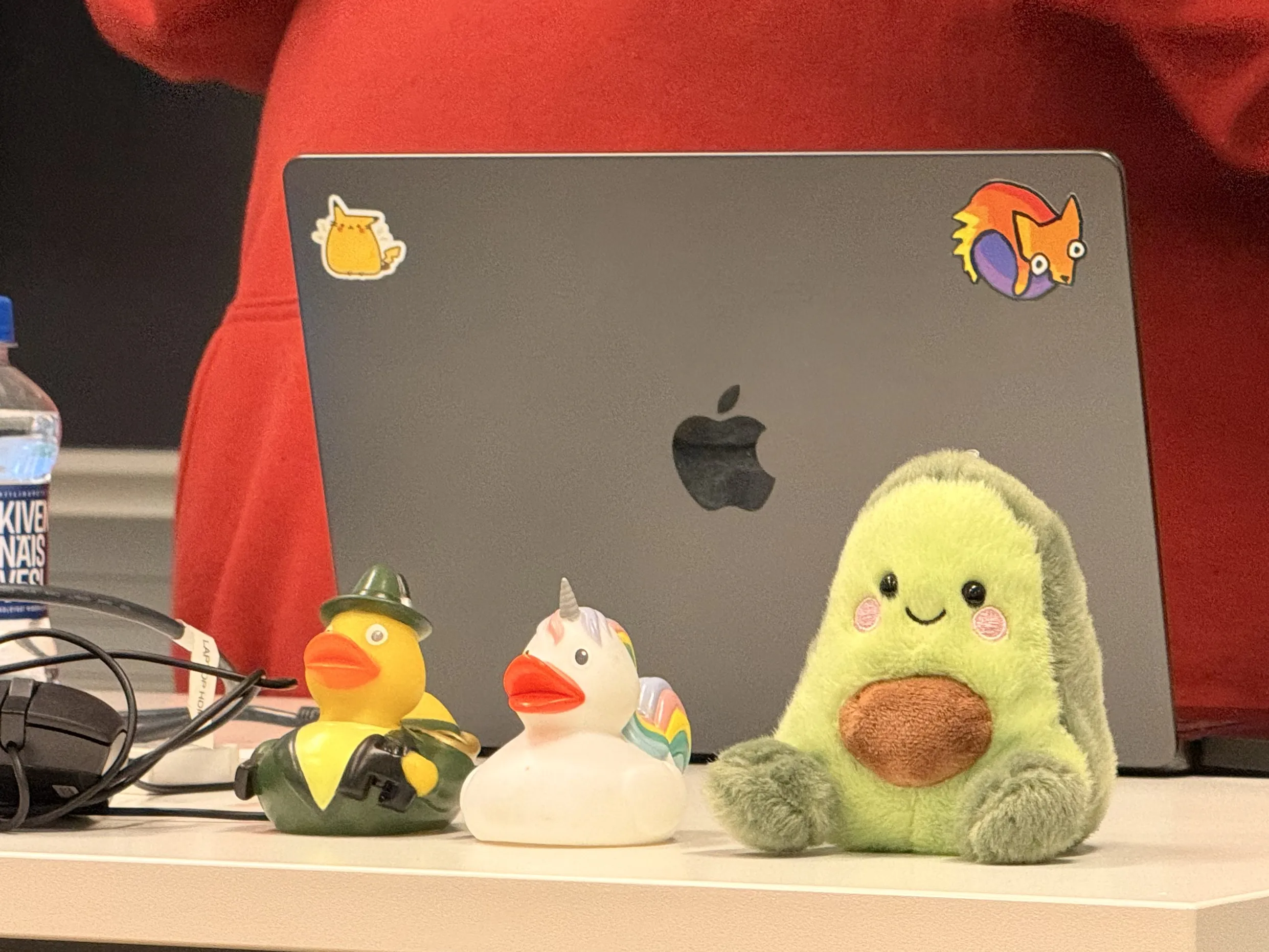 Two rubber ducks: one dressed as a hunter and another is a unicorn and an avocado plush in front of a laptop that has a quirky Pikachu and Firefox stickers on its cover.