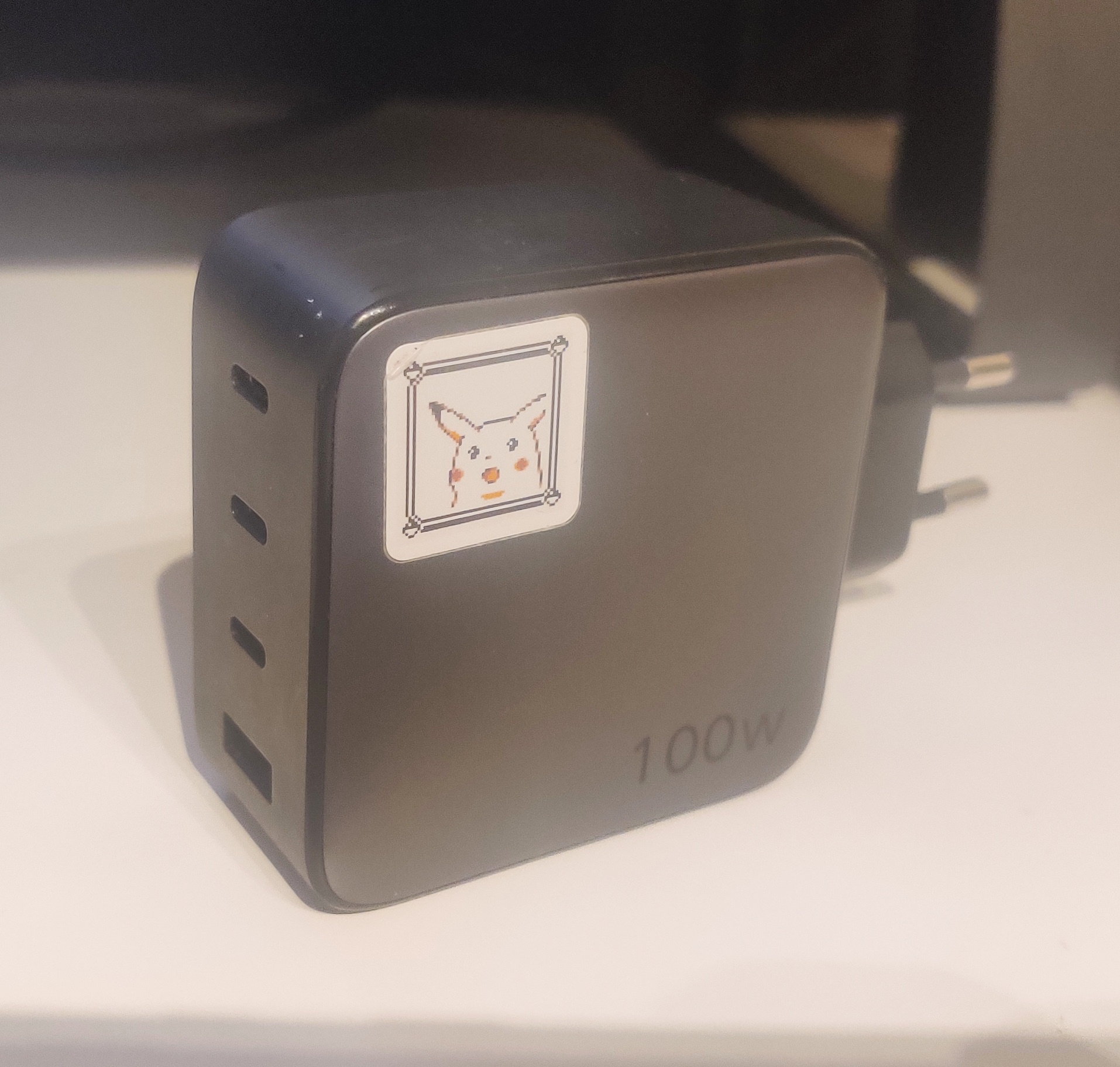 A dark grey 100W charger with 3 USB-C ports and 1 USB-A port. A surprised Pikachu sticker in one corner. 