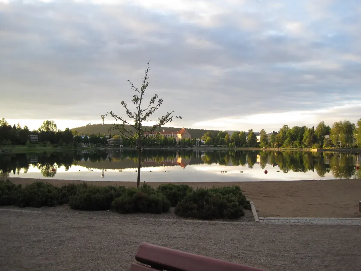 A sunset, a large pond and a small beach. The pond is surrounded by trees on the opposite side and there’s a couple of buildings peeking from behind the trees. 