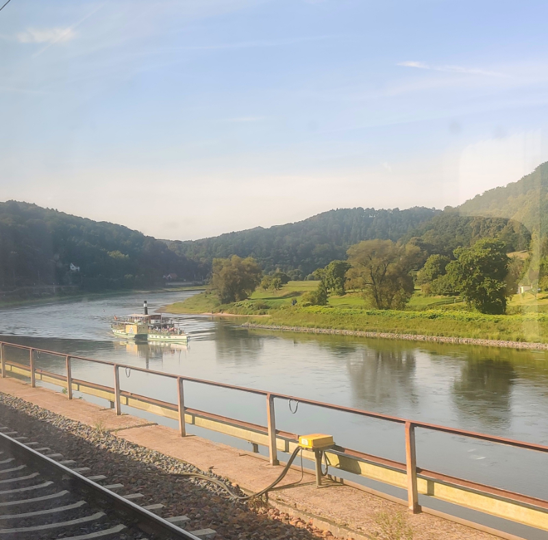 A photo taken from the train with a river and a river boat and on the other side of the river, a patch of land with trees and hills rising from beyond that land 