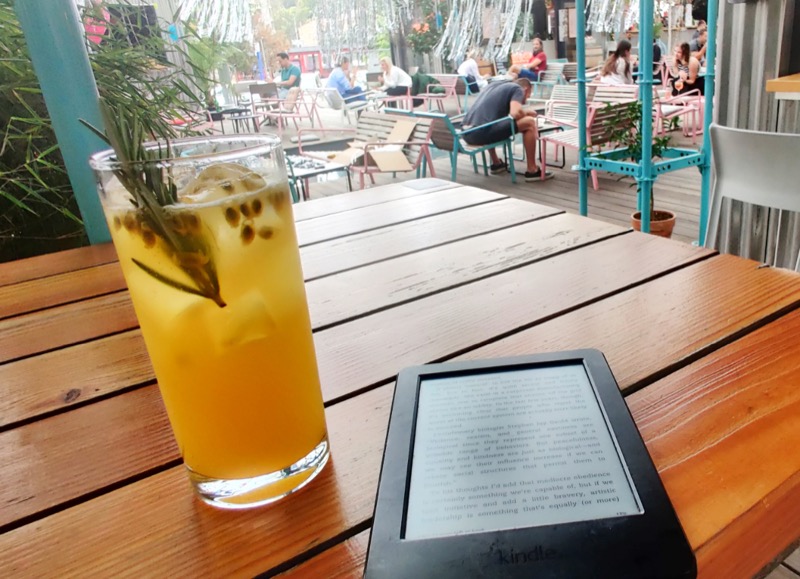 A yellow cocktail on a glass, a Kindle with a book open on it and on the background, some people sitting on chairs enjoying life. 