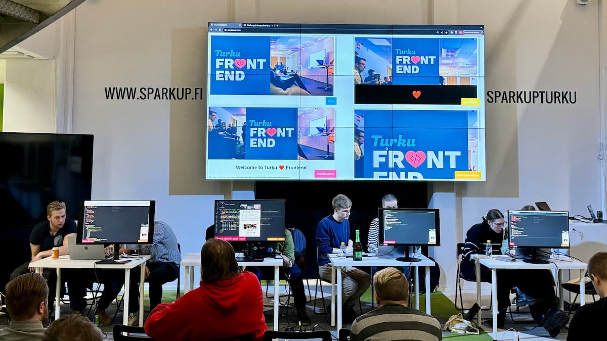 Four teams sitting behind desks. The desks have displays that show their code editors. Behind them, a large screen that shows four previews of Turku Frontend websites the teams are building. 