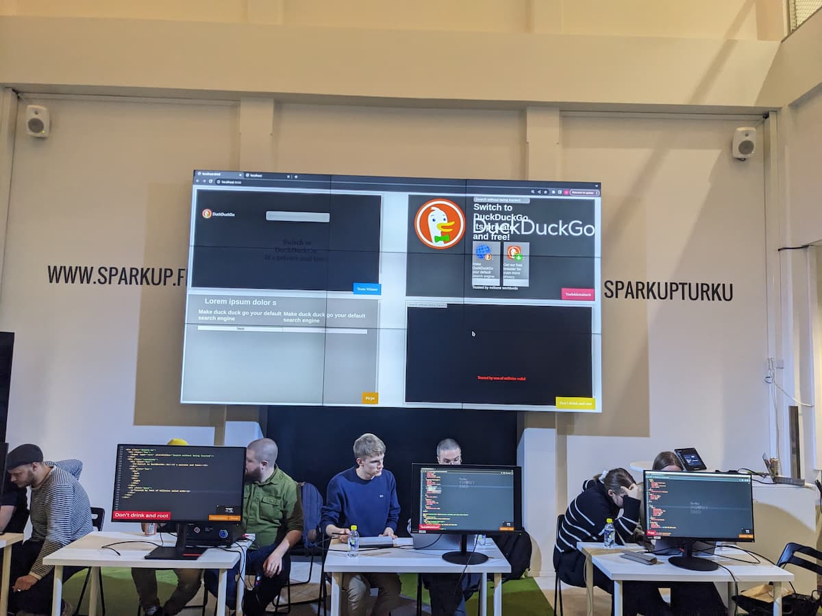 Four teams sitting behind desks. The desks have displays that show their code editors. Behind them, a large screen that shows four previews of DuckDuckGo’s front pages the teams are replicating. 