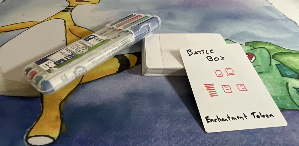 A stack of dry erase cards, four markers (black, red, green and blue) and a single card with text Battle Box - Enchanment Token and a poor drawing of a Magic battlefield with a deck and four cards. 