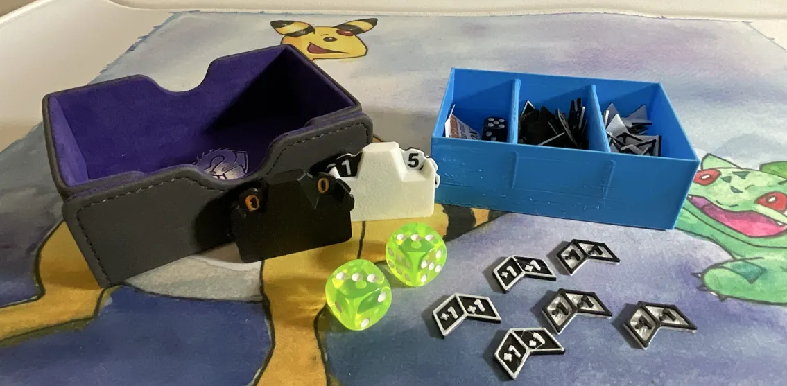 A collection of small tokens next to their box. Two double-digit life counters, two bright green six sided dice, six black and white 3D printed chevron shaped counters and a blue box full of those same counters. 