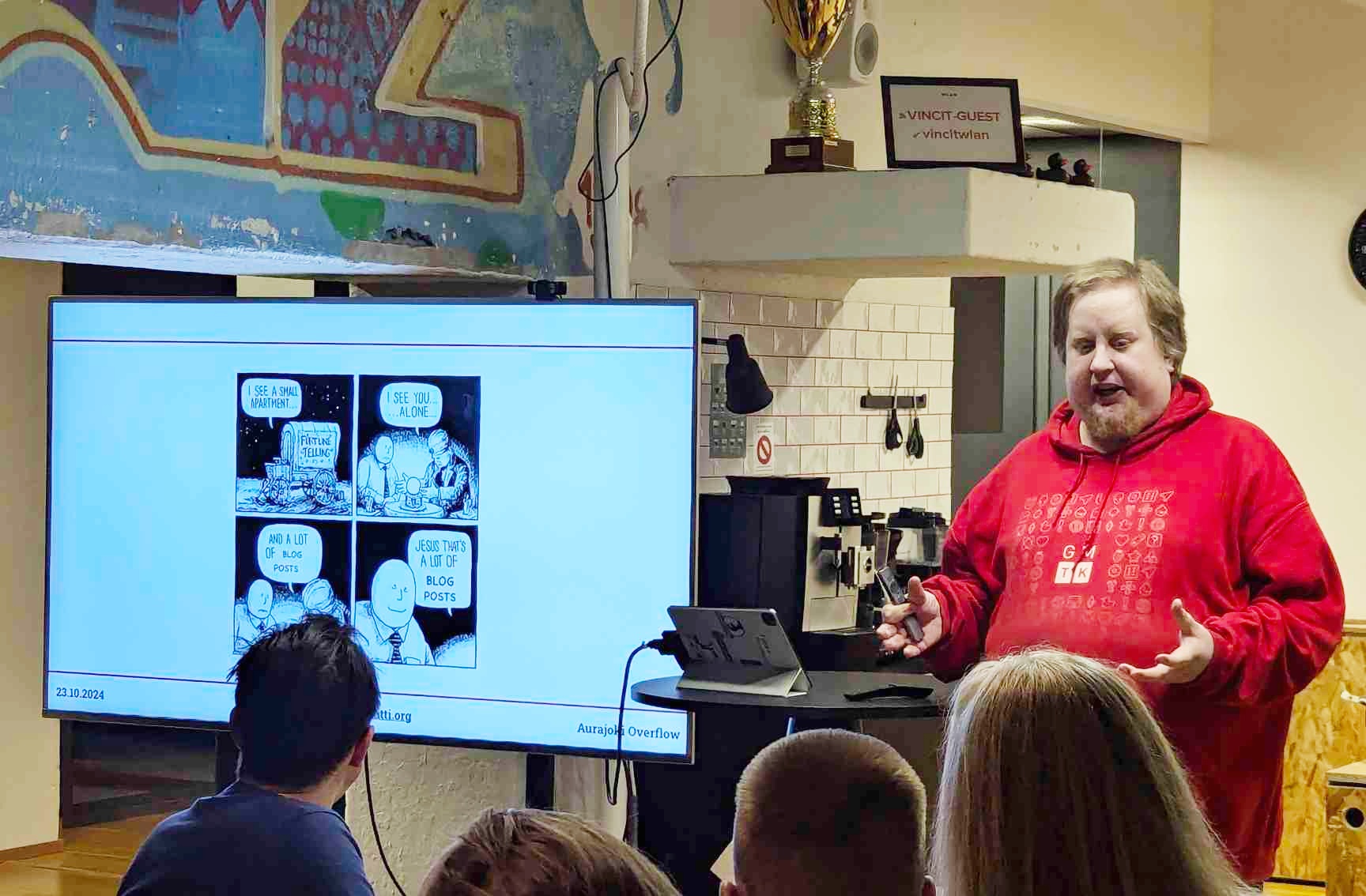 Juhis wearing a red hoodie stands in front of an audience that is partially visible in the photo. A large TV is next to him showing a four pane comic meme with illegible text. 