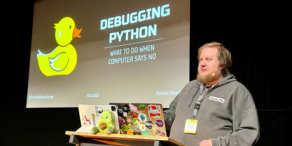 Juhis standing behind a speaker podium on stage with a large screen behind him with a slide that reads Debugging Python, what to do when computer says no