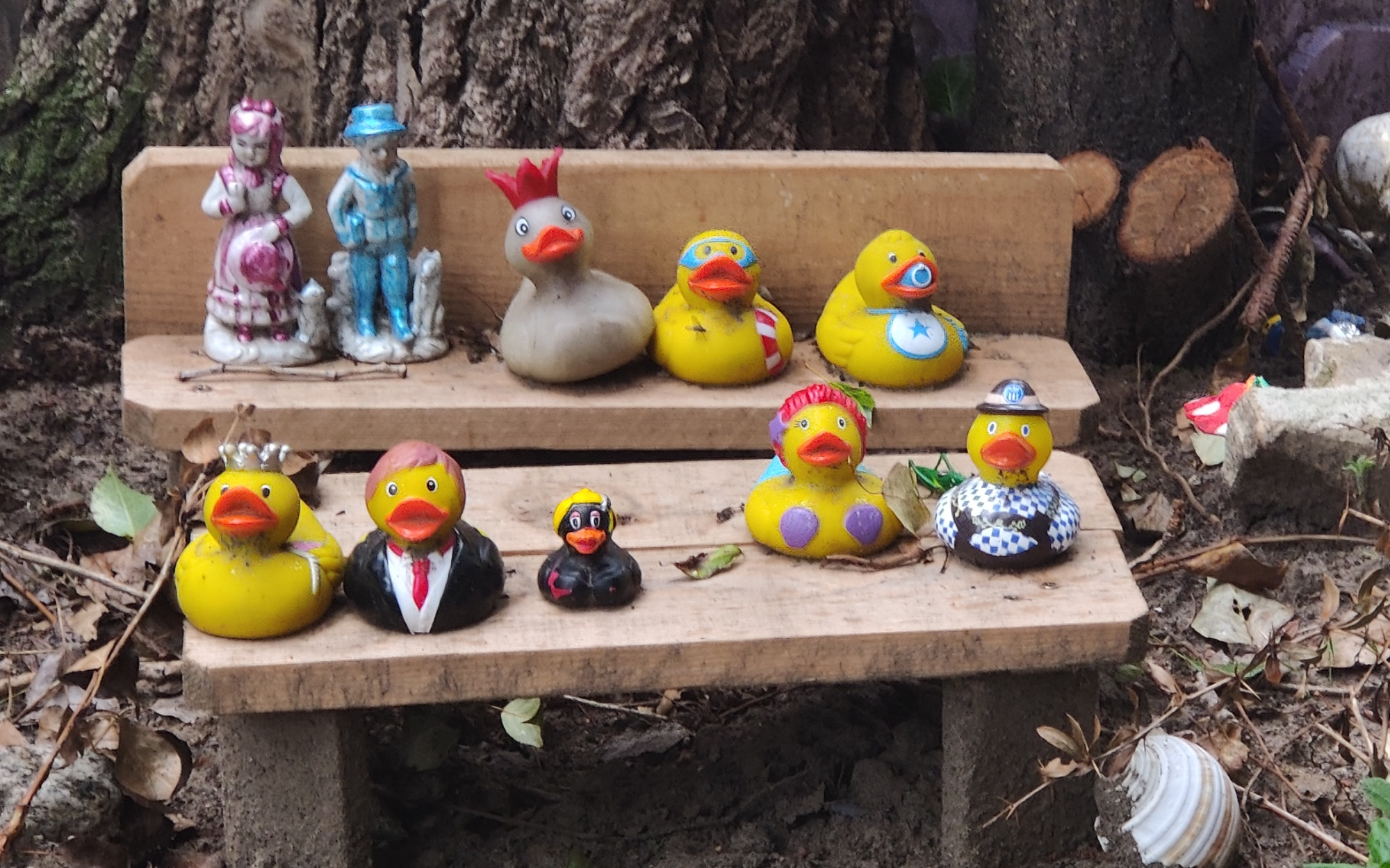 A small bench at the foot of a tree, with two rows of differently themed rubber ducks.