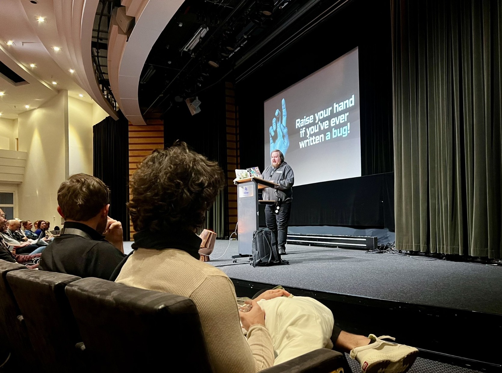 Juhis talking on a stage with a slide that reads "Raise your hand if you've ever written a bug". A couple of people in the foreground listening to a talk.