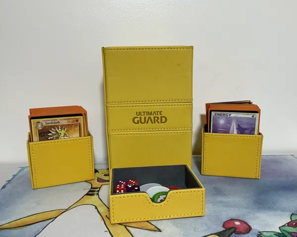 Yellow Ultimate Guard deck box with two of its smaller containers next to it. One has a stack of cards, showing Sandslash and another has a stack of cards showing a Psychic energy. In front of them, a token container showing a few dice and special condition markers.